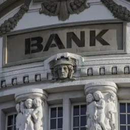 Fasada banku z kamienną tablicą z napisem 'BANK', ozdobiona rzeźbami twarzy i postaci dzieci, widziana z dołu w pochmurny dzień.