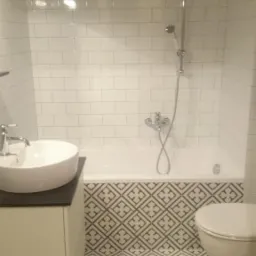 Wykończona łazienka z białą armaturą, geometrycznymi płytkami podłogowymi i białymi płytkami ściennymi ułożonymi w cegiełkę, oświetlona lampą punktową.