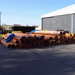 Pomarańczowe rury kanalizacyjne różnych średnic ułożone na placu, z budynkiem magazynowym w tle i fragmentem nieba. Widoczny zwój niebieskiej rury.