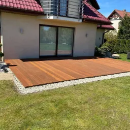 Nowoczesny taras z drewna egzotycznego w ogrodzie, wykończony białym kamieniem, przylegający do domu z dużymi przesuwnymi drzwiami tarasowymi.