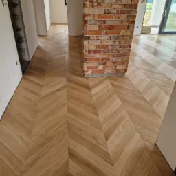 Ułożona podłoga z drewnianych paneli w jodełkę, widoczny fragment ściany z cegły, surowy stan wykończenia wnętrza.