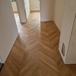 Ułożona podłoga z parkietu w jodełkę francuską, widok z perspektywy wejścia do pomieszczenia, fragmenty ścian w stanie surowym.