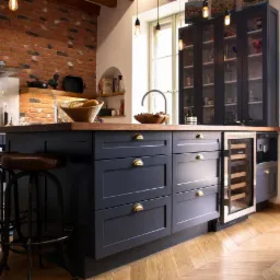 Wnętrze kuchni z wyspą w kolorze granatowym, drewnianym blatem i złotymi uchwytami, na podłodze parkiet ułożony w jodełkę, w tle ściana z czerwonej cegły i przeszklona witryna.