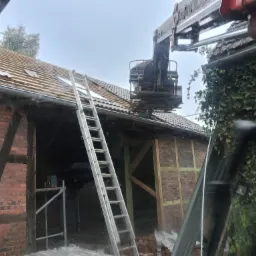 Częściowo zdjęty dach z ceglanej stodoły, widoczna drabina i dźwig. Prace dekarskie w toku, zieleń na ścianie budynku. Pochmurne niebo w tle.