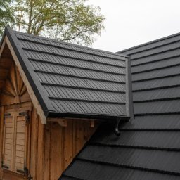 Dekarstwo Polskie - Czarny dach z blachy na drewnianym domu z oknem. Widoczne drzewa w tle. Dach pokryty równymi rzędami blachy, tworzącymi wzór przypominający dachówkę.