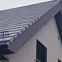 Szary dach z dachówki ceramicznej, z zamontowanym systemem odladzania i orynnowaniem, na tle szarej elewacji domu z wentylacją.