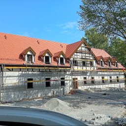 Dekarstwo Polskie - Budynek w trakcie renowacji z rusztowaniami, nowym czerwonym dachem i elementami konstrukcji szkieletowej. Widoczny stos kamieni i pracownik. Słoneczny dzień.
