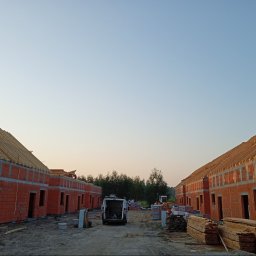 Dekarstwo Polskie - Wczesny etap budowy szeregu domów z czerwonej cegły. Widoczna więźba dachowa, materiały budowlane i pojazd dostawczy. Tło: drzewa i niebo o świcie.