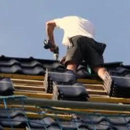 Dekarz mocuje ciemną dachówkę do drewnianej konstrukcji dachu w słoneczny dzień. Widoczne narzędzia i fragment rusztowania.