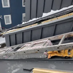 Szary dach z blachy na rąbek stojący w trakcie prac, widoczna drabina zabezpieczona taśmą, narzędzia dekarskie i fragment elewacji budynku z oknami.