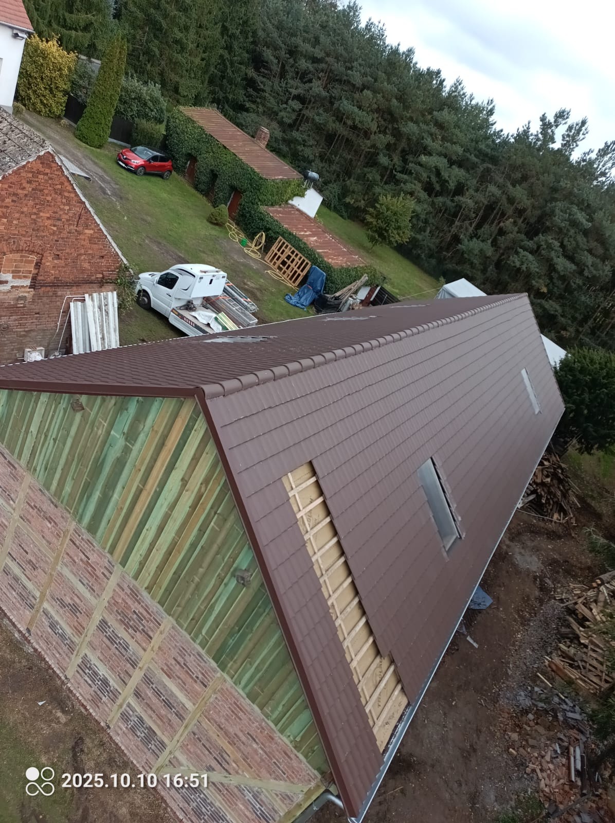 Brązowy dach z dachówki ceramicznej w trakcie montażu, widoczne fragmenty konstrukcji i okno dachowe. W tle zieleń drzew i budynki.