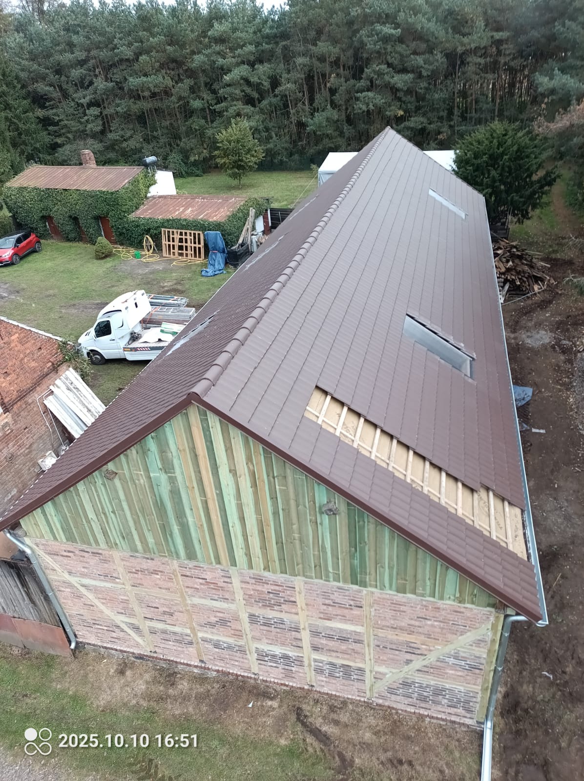 Brązowy dach z lukarnami w trakcie krycia. Widoczna więźba dachowa i fragment starego dachu. Na posesji zaparkowany biały bus. Ujęcie z góry.