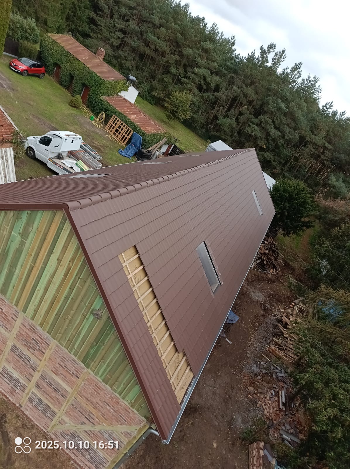Brązowy dach z lukarnami w trakcie krycia, widoczna więźba i fragment muru. Na posesji biały samochód dostawczy i sterta drewna. Ujęcie z góry, lekko obrócone.