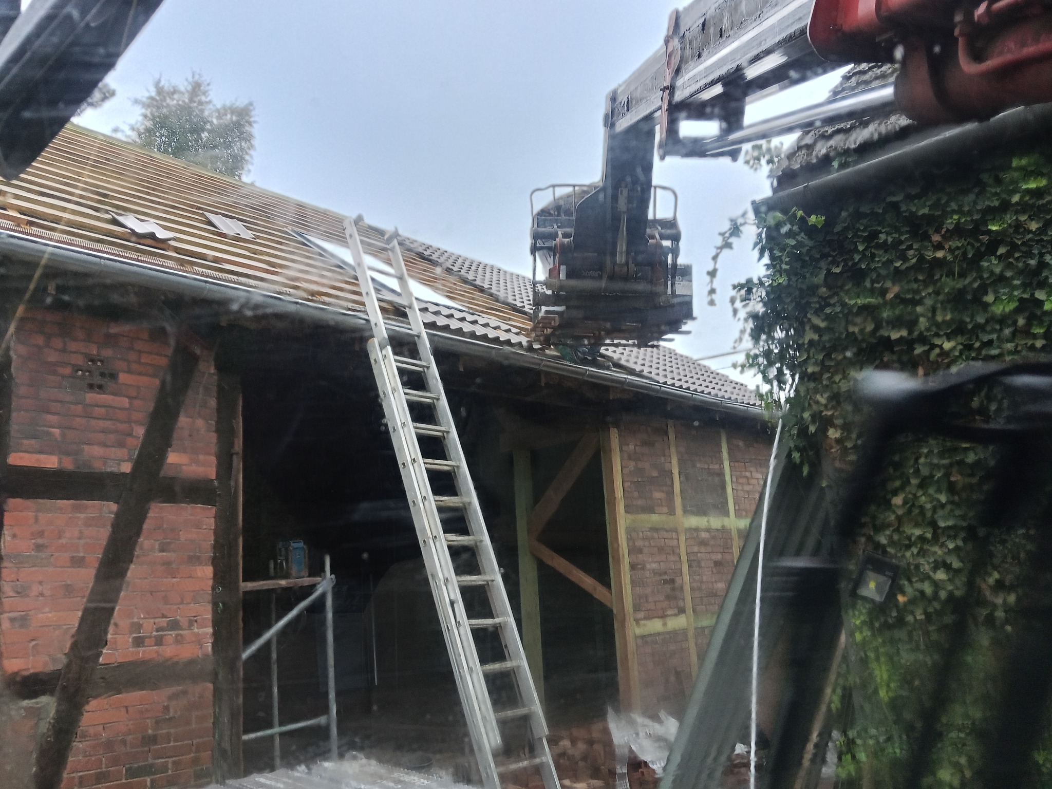 Częściowo zdjęty dach z ceglanej stodoły, widoczna drabina i dźwig. Prace dekarskie w toku, zieleń na ścianie budynku. Pochmurne niebo w tle.