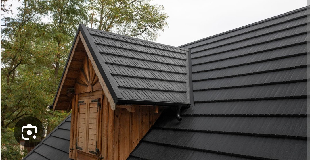 Czarny dach z blachy na drewnianym domu z oknem. Widoczne drzewa w tle. Dach pokryty równymi rzędami blachy, tworzącymi wzór przypominający dachówkę.