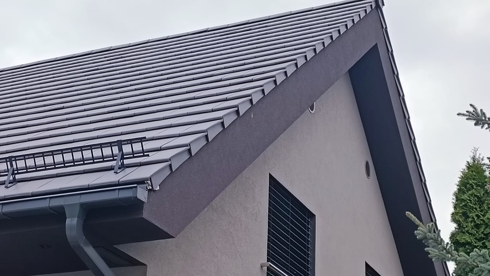 Szary dach z dachówki ceramicznej, z zamontowanym systemem odladzania i orynnowaniem, na tle szarej elewacji domu z wentylacją.