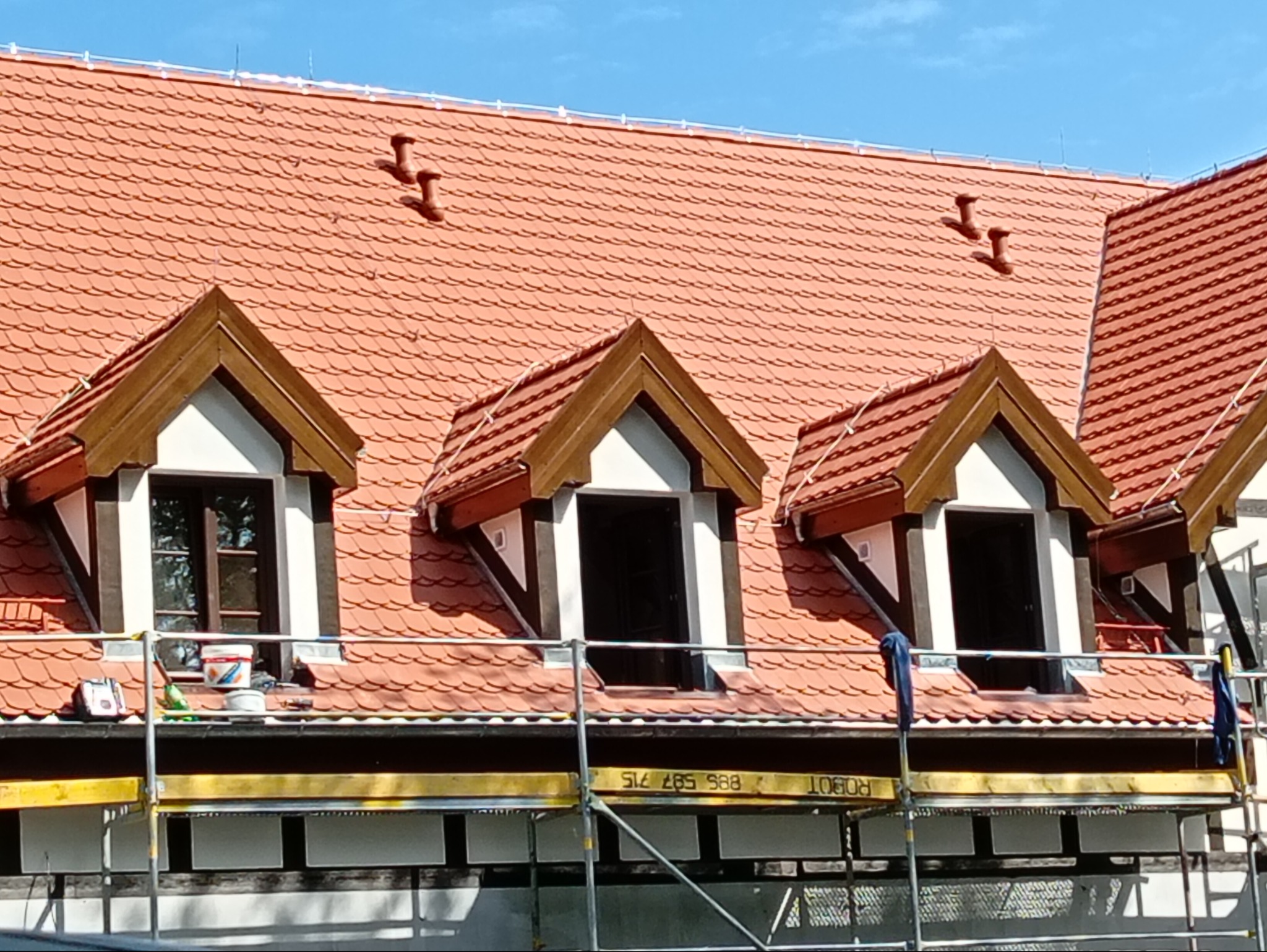 Czerwona dachówka karpiówka na dachu z lukarnami, widoczne rusztowanie i akcesoria malarskie. Prace dekarskie w trakcie renowacji.