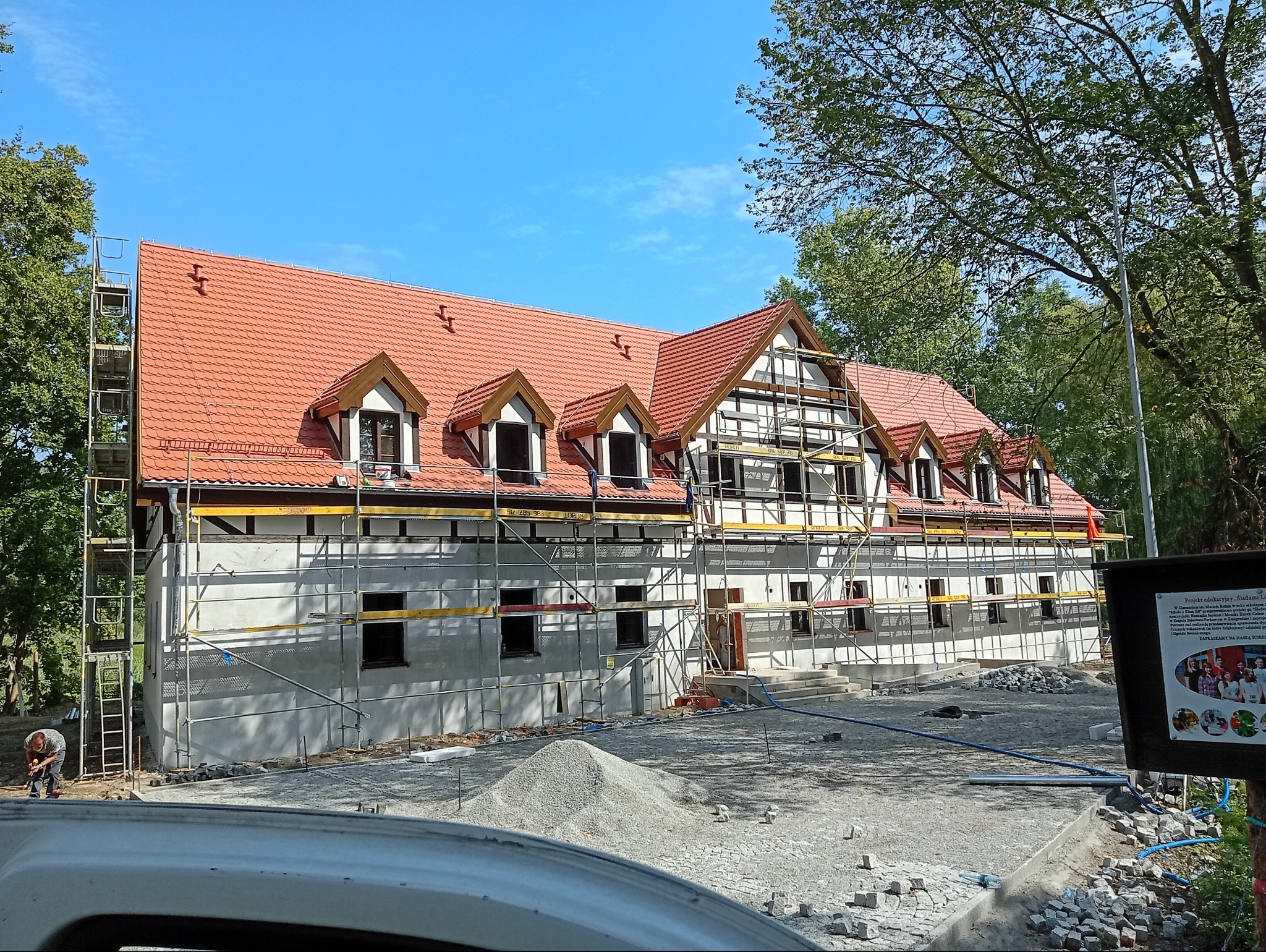 Budynek w trakcie renowacji z rusztowaniami, nowym czerwonym dachem i elementami konstrukcji szkieletowej. Widoczny stos kamieni i pracownik. Słoneczny dzień.