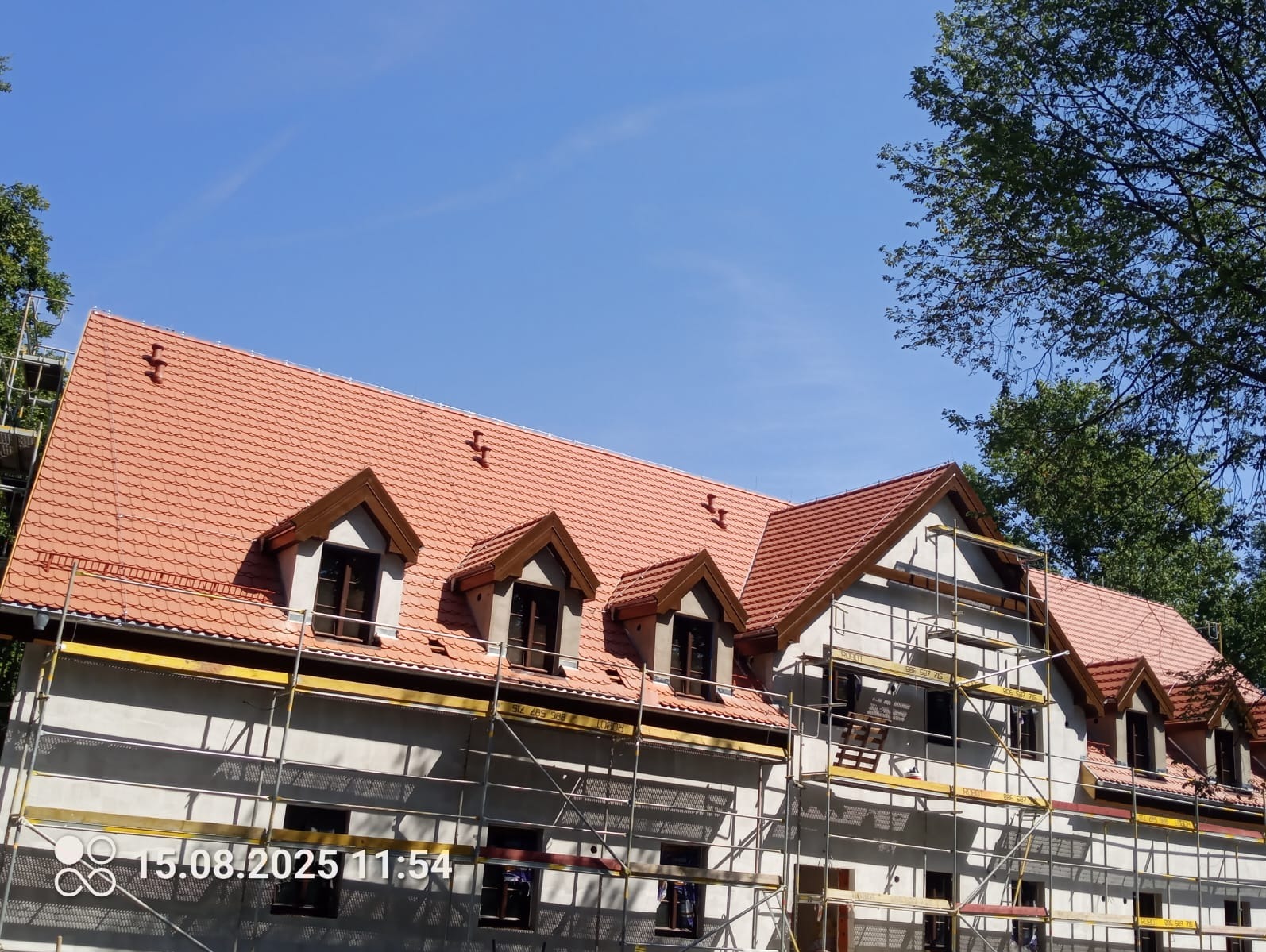 Budynek w trakcie remontu dachu z nową, czerwoną dachówką. Widoczne rusztowania i okna dachowe. Błękitne niebo w tle. Ujęcie z dołu, podkreślające rozległość prac.