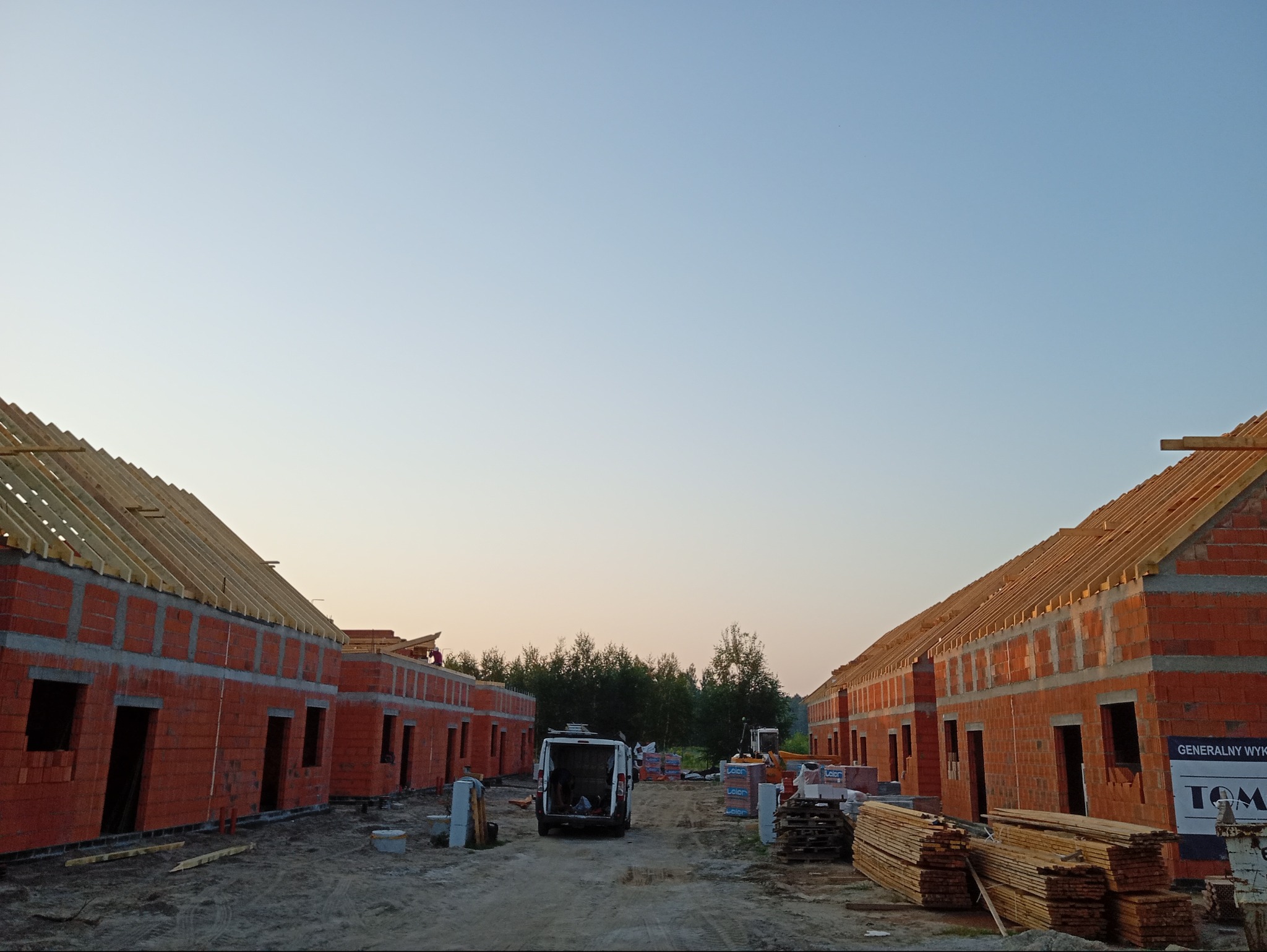 Wczesny etap budowy szeregu domów z czerwonej cegły. Widoczna więźba dachowa, materiały budowlane i pojazd dostawczy. Tło: drzewa i niebo o świcie.