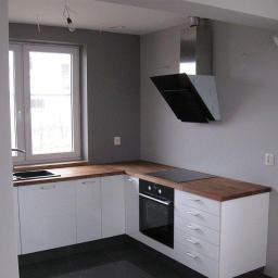 Zakład Stolarski KASARO - Nowoczesna kuchnia w szaro-białej tonacji z drewnianym blatem, czarną płytą grzewczą i okapem. Widoczne białe szafki i zlewozmywak przy oknie. Minimalistyczny design.