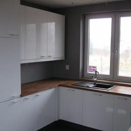 Zakład Stolarski KASARO - Nowoczesna kuchnia z białymi szafkami i drewnianym blatem pod oknem. Widoczny zlew i bateria. Minimalistyczny design, szare ściany.