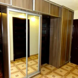 Brązowa szafa wnękowa z lustrem i przesuwnymi drzwiami, z nadstawką, na tle wzorzystej podłogi. Widoczne drzwi do innego pomieszczenia.