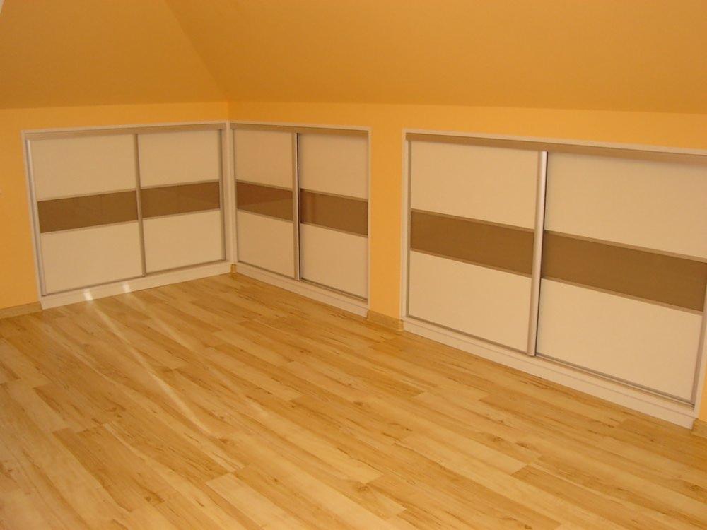 Dwa wbudowane, przesuwne systemy szaf wnękowych z beżowymi i białymi frontami, dopasowane do skośnego sufitu na poddaszu, widok na jasną drewnianą podłogę.