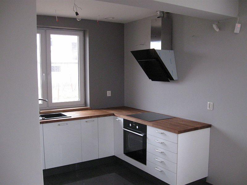 Nowoczesna kuchnia w szaro-białej tonacji z drewnianym blatem, czarną płytą grzewczą i okapem. Widoczne białe szafki i zlewozmywak przy oknie. Minimalistyczny design.