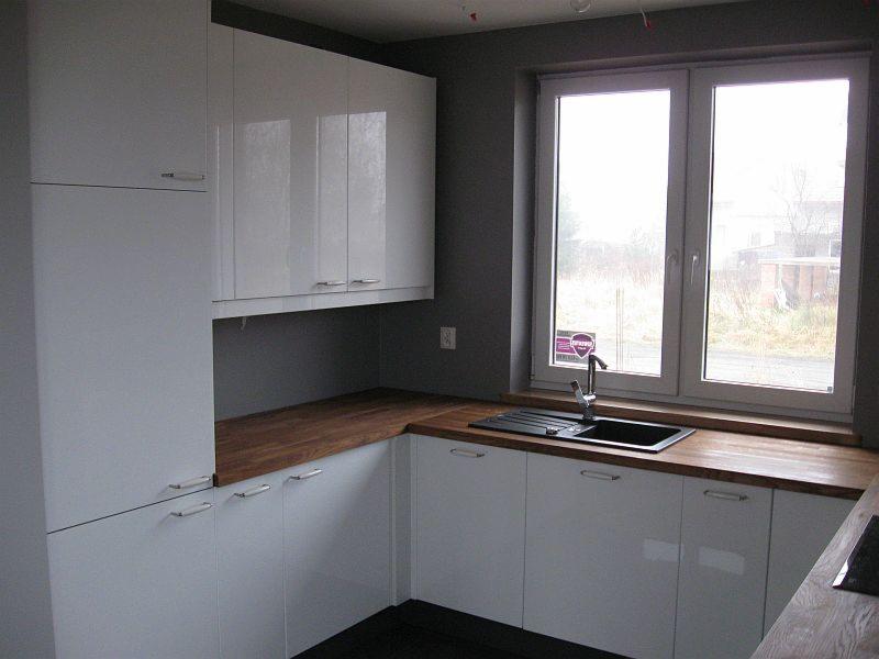 Nowoczesna kuchnia z białymi szafkami i drewnianym blatem pod oknem. Widoczny zlew i bateria. Minimalistyczny design, szare ściany.