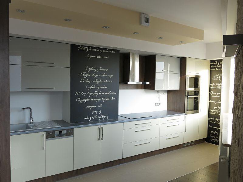 Nowoczesna zabudowa kuchenna w odcieniach bieli i szarości, z tablicą z przepisem na ścianie, oświetleniem punktowym i drewnianymi akcentami.