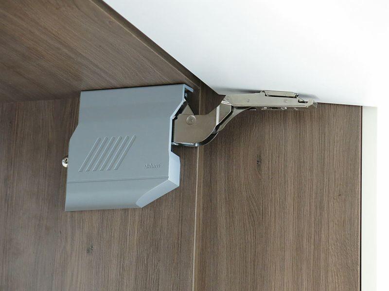 Szary mechanizm podnoszenia Blum zamontowany w szafce z drewnopodobnymi drzwiami. Widoczne mocowanie do korpusu i ramię do frontu.