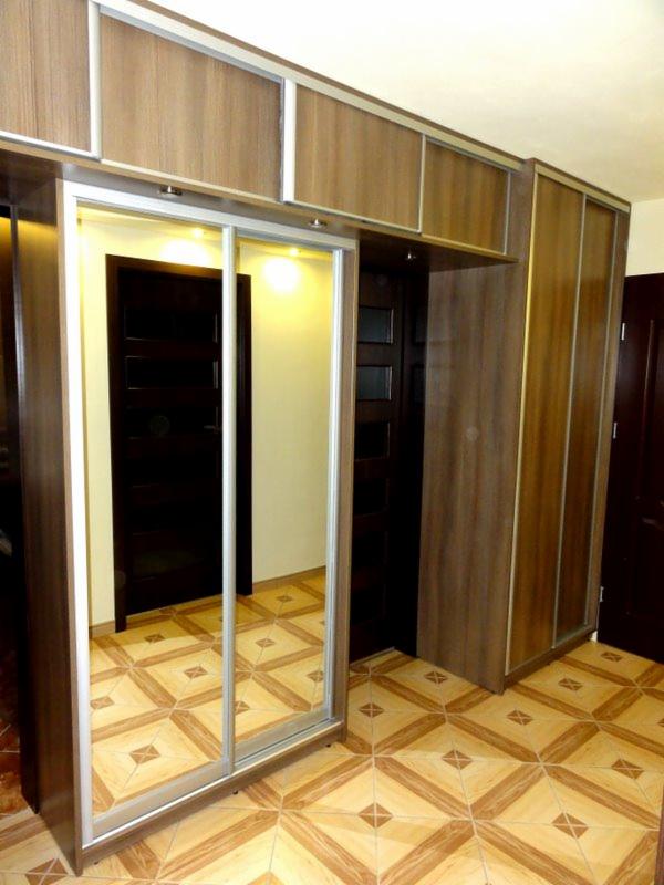 Brązowa szafa wnękowa z lustrem i przesuwnymi drzwiami, z nadstawką, na tle wzorzystej podłogi. Widoczne drzwi do innego pomieszczenia.