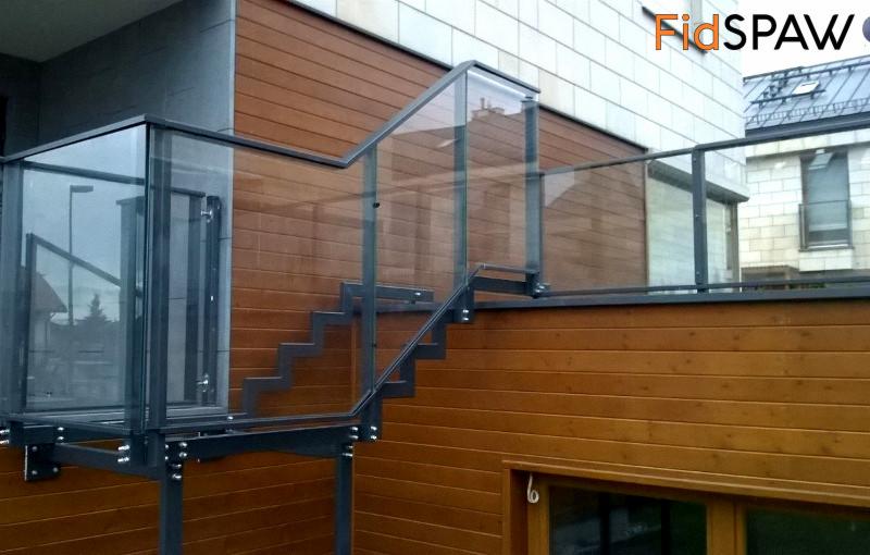 Metalowa konstrukcja schodów zewnętrznych z szklanymi balustradami, zamontowana przy drewnianej elewacji budynku. Widoczne detale połączeń śrubowych.