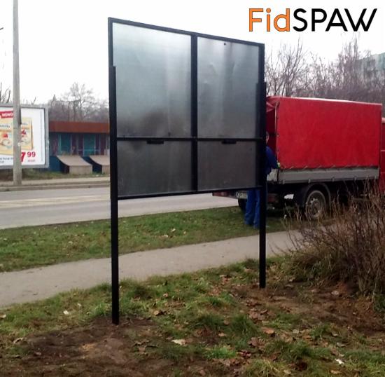 Metalowa konstrukcja tablicy reklamowej z czarną ramą, stojąca na trawie przy chodniku, z widocznym fragmentem czerwonej ciężarówki w tle.