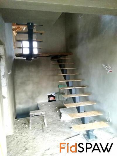 Wnętrze w trakcie remontu, z widocznymi schodami stalowymi o drewnianych stopniach, zamontowanymi w surowym stanie. Widoczne ślady prac budowlanych.