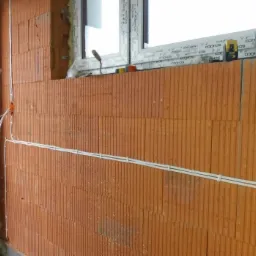 Czerwona ściana z pustaków ceramicznych z częściowo poprowadzonymi przewodami elektrycznymi w białych listwach, widoczna puszka instalacyjna, okno z narzędziami na parapecie i laserowy poziomica.