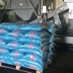 Stos worków z ekogroszkiem na palecie, z czerwonym wózkiem widłowym i metalową maszyną pakującą w tle.