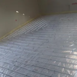 Ułożone rury ogrzewania podłogowego na folii, widoczne złącza i siatka zbrojeniowa, przygotowane do wylania posadzki w jasnym pomieszczeniu.