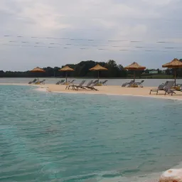 Sztuczna plaża z turkusową wodą, leżakami i strzechami, otoczona zielenią i jeziorem, widok z poziomu wody.