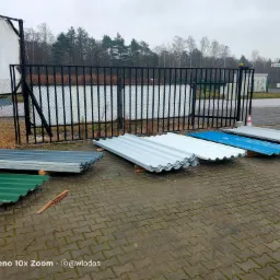 Stosy pofalowanych arkuszy blachy w różnych kolorach (zielony, brązowy, srebrny, niebieski) ułożone na placu wyłożonym kostką, z widocznym ogrodzeniem i stacją benzynową w tle.