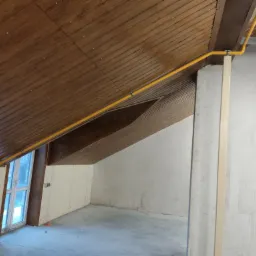 Żółta instalacja gazowa poprowadzona wzdłuż drewnianego sufitu poddasza, z widocznymi elementami mocującymi i zaworem przy oknie, w surowym, nieukończonym wnętrzu.