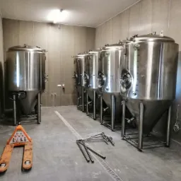 Rząd błyszczących, stożkowych zbiorników ze stali nierdzewnej w sterylnym pomieszczeniu, z pomarańczowym wózkiem paletowym i rurami na podłodze, sugerujący etap instalacji.