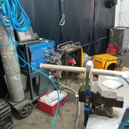 Pro-Weld Industry Service Sp. z o.o. - Stanowisko robocze z niebieskim urządzeniem spawalniczym Orbitallum, butlą gazową na kółkach i rurą zamocowaną w imadle, przygotowaną do spawania orbitalnego, w tle nagrzewnica Master i czarna kotara.
