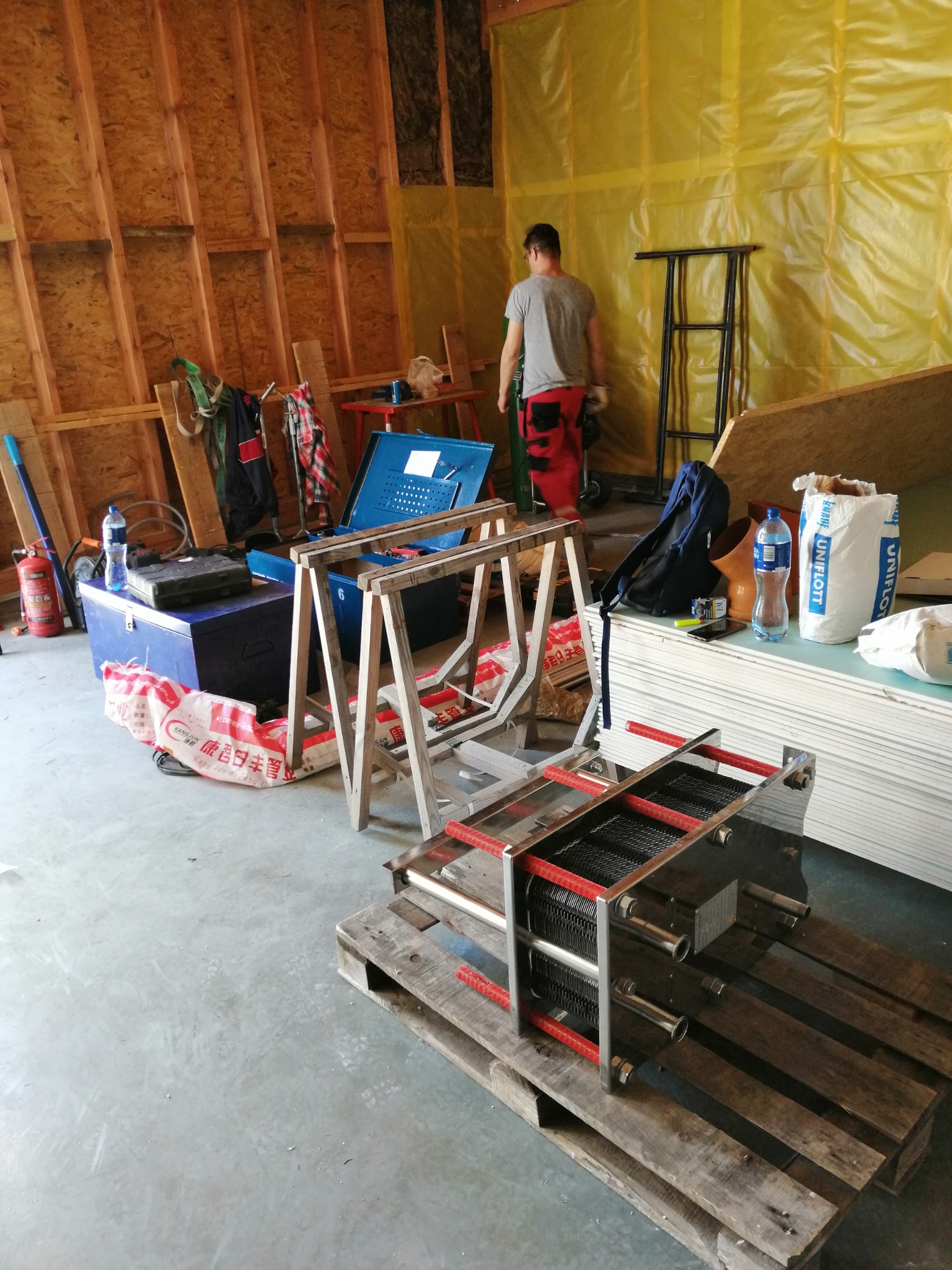 Wymiennik ciepła na palecie w surowym wnętrzu, widoczne narzędzia, skrzynki i materiały budowlane, mężczyzna w tle