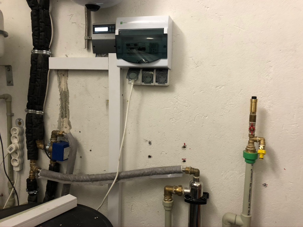 Instalacja z rurami, zaworami i osprzętem elektrycznym na kremowej, chropowatej ścianie w pomieszczeniu technicznym. Widoczne elementy systemu grzewczego i zasilania.