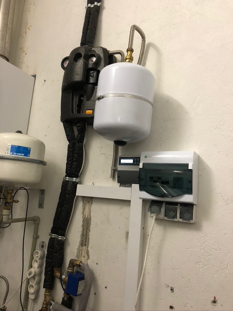 Instalacja centralnego ogrzewania z czarnymi rurami w izolacji, białym zbiornikiem wyrównawczym i skrzynką elektryczną na ścianie o nierównej fakturze.