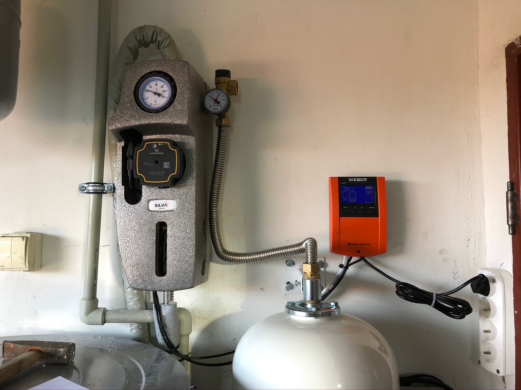 Instalacja zasilana energią słoneczną: szary panel sterowania z manometrami, pomarańczowy regulator temperatury Weber, biały zbiornik oraz rury w jasnym pomieszczeniu.