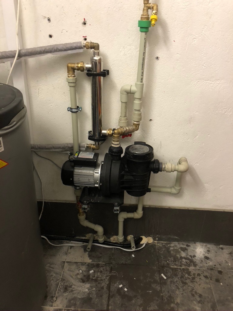 Instalacja solarna z pompą obiegową, filtrem i rurami z tworzywa sztucznego, zamontowana przy białej ścianie z widocznymi śladami po wierceniu i nieestetyczną podłogą.