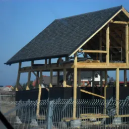 Konstrukcja domu w trakcie budowy z drewnianym szkieletem i czarnym dachem z gontu bitumicznego, widoczna przez metalowe ogrodzenie.